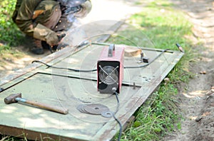 Old man welder in brown uniform, welding mask and welders leathers, weld metal door with arc welding machine outdoors