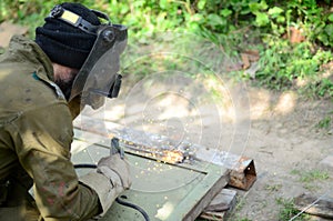 Old man welder in brown uniform, welding mask and welders leathers, weld metal door with arc welding machine outdoors