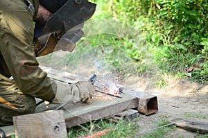 Old man welder in brown uniform, welding mask and welders leathers, weld metal door with arc welding machine outdoors