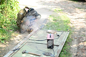 Old man welder in brown uniform, welding mask and welders leathers, weld metal door with arc welding machine outdoors