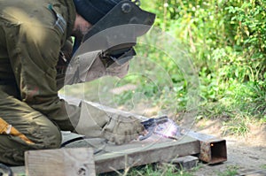 Old man welder in brown uniform, welding mask and welders leathers, weld metal door with arc welding machine outdoors