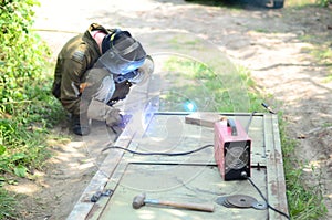 Old man welder in brown uniform, welding mask and welders leathers, weld metal door with arc welding machine outdoors