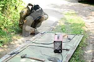 Old man welder in brown uniform, welding mask and welders leathers, weld metal door with arc welding machine outdoors