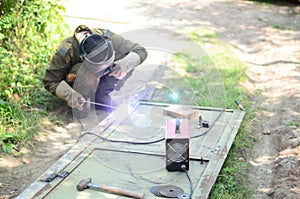 Old man welder in brown uniform, welding mask and welders leathers, weld metal door with arc welding machine outdoors