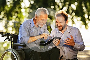 Old man in a wheelchair and a man looking at something in a tablet walking in the park. They are happy