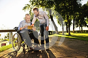 Old man in a wheelchair and a man looking at something in a tablet walking in the park. They are happy