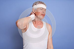 Old man in white t shirt holding neck in pain isolated over blue background, touches her neck, has injury during sport training,