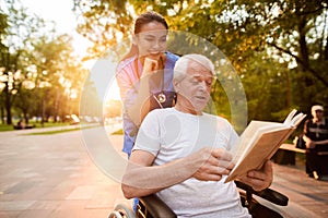 The old man who is sitting in a wheelchair is reading a book. A nurse behind the old man and also looking at the book