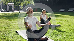 Old Man And Woman Practicing Yoga In Park. They Sit On Rugs On Ground. Their Eyes Are Closed.