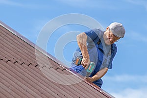 Old man working at heat on a roof of a house with electric screwdriver, wearing no safety devices, work clothing, blue overall, da