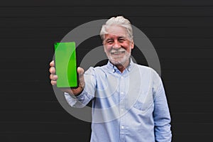 Old and mature man holding his smartphone showing it at the camera with the green screen - smiling happy adult portrait using