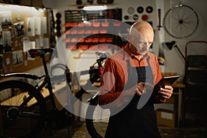 Old mechanic using digital tablet repairing a bicycle in a workshop or garage.