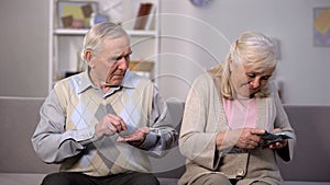Old man counting coins looking at happy senior woman holding dollars, wealth