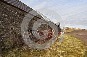 Old Metal Farm gates stacked against a wall of a Farm Stone built Outbuilding along with other Farm Debris.