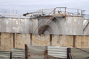 Old metal industrial storage tanks with rusty inspection ladders and valves surrounded by a corrugated steel fence and wall