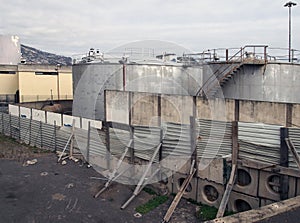 Old metal industrial storage tanks with rusty inspection ladders and valves surrounded by a corrugated steel fence and wall