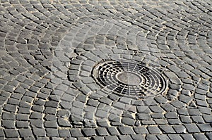 Old metal sewer hatch in the middle of the road, laid out of a smooth paving ston