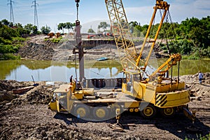 Old mobile crane truck with pneumatic hammer on building site. Bridge foundation made of metal piles on river coast