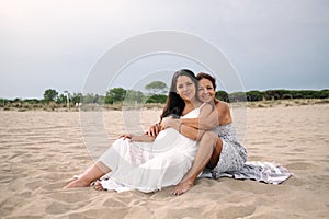 Old mother and pregnant adult daughter are seen hugging and smiling on a beach with white dress