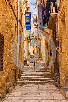 Old narrow medieval street with yellow buildings with colorful balconies in town Singlea, Valletta, Malta