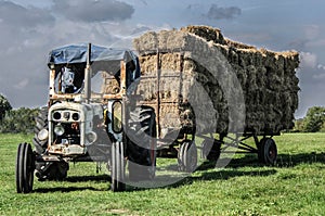 Old neglected blue tractor pulling a triler of hay in a field at harvest time