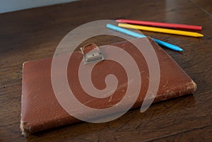 Old notebook with leather cover on a wooden table and colorful pencils