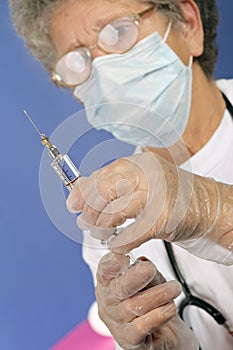 Old nurse with protective mask and gloves prepares injection of vaccine or medicine for disease prevention with glass syringe and