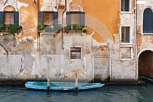 Old orange building with open windows and plants and a blue boat in the Venice canal, Italy
