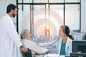 Old patient man sitting in wheelchair while young female doctor using stethoscope to check his heartbeat, elderly health checkup,