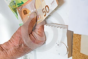 Old pensioner man holding banknotes in hand in front of a heating thermostat in an apartment, Germany