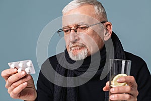 Old pensive sick man with gray hair and beard in eyeglasses and scarf thoughtfully looking on pills holding glass of
