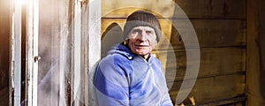 Old person in knitted cap seated on wooden bench against wall with window.