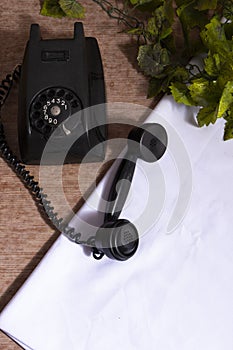 Old phone on wooden table with white tablecloth and artificial green plants. Black antique on a table