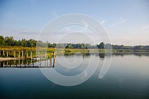 Old pier with a boat on the lake with reeds, trees and fishermen. The sky is reflected in the water surface. Landscape. Morning