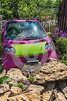 Old pink and green planted car used as a decoration and flower pot in a garden. A watercourse is installed in front