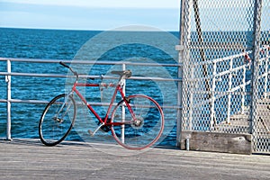 An old red bicycle leaning against and chained to a fence and hand rail on a wooden pier with the blue ocean in the background