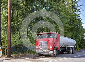 Old red big rig cabover semi truck with tank trailer standing on the road shoulder on the green trees background