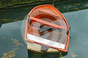 An old red boat filled with water moored with a rope in the harbor. Sea water with seaweed and raindrops.