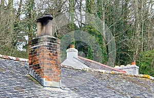 Old red brick chimney stack on a slate roofed building, with another old roof behind it and trees in the background