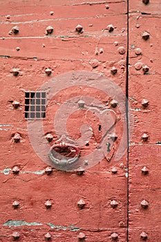 An old red door of medieval style with metallic rivets, heart shaped keyhole plate, cast iron knocker and wire mesh spyhole