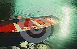 An old red fishing boat with oars floats by the riverbank on a calm summer day, reflecting the bright sunbeams on its surface. The