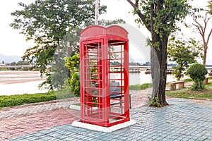 Old Red phone booth located on side to river with evening atmosphere. Green trees, bridge over river background.