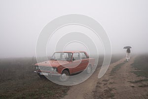 Old red retro car stands in fog near the dirt road, girl in white coat walks under umbrella on the background