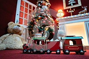 Old, red toy metal train, in background an open fireplace with a Christmas tree, gifts underneath, a large teddy bear, dark red,