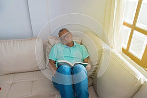 An old retired man reading a book on his couch by the window