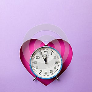 Old retro analog blue alarm clock in pink heart background on purple background. The clock starts from five minutes to twelve o`
