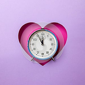 Old retro analog blue alarm clock in pink heart background on purple background. The clock starts from five minutes to twelve o`