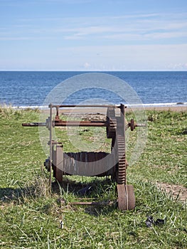 An Old rusting Manual Boat Winch used for hauling in Fishing Boats lies abandoned on the Grassy Bank behind the Beach.