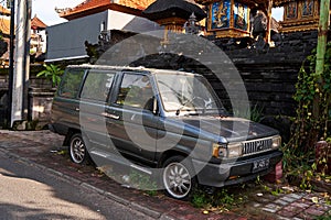 An old, rusty, abandoned car is standing in the grass on the side of the road