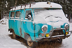 An old rusty ambulance van stands in a snow-covered clearing on a cloudy winter day